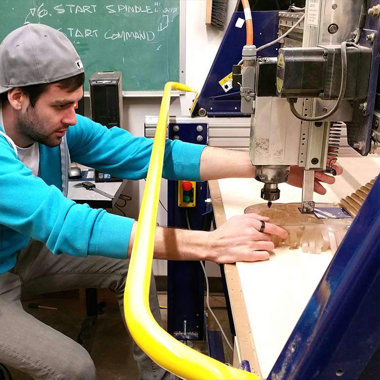 A male student uses a CNC machine to carve a piece of furniture in the Think It Make It Lab.