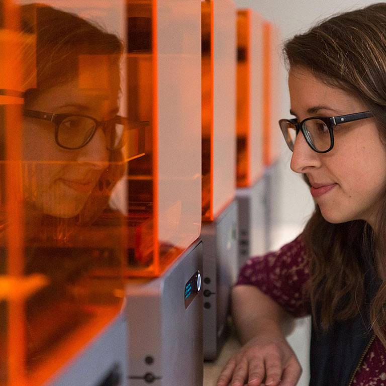 A female student watches a 3D printer create her design in the Think It Make It Lab.