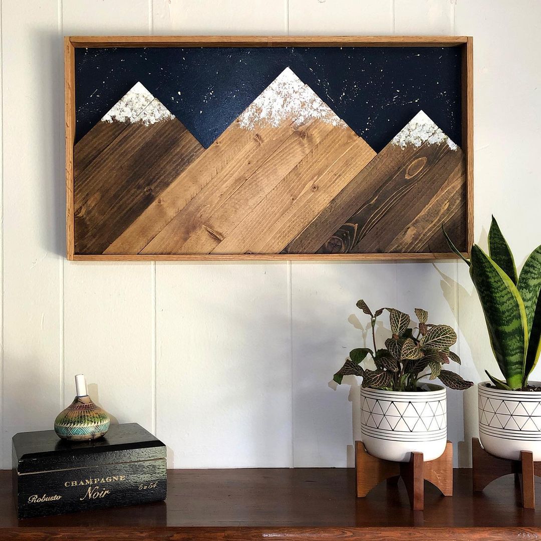 Three peaks with white paint for snow caps in a rustic wooden frame, hanging on a while wall with small house plants underneath