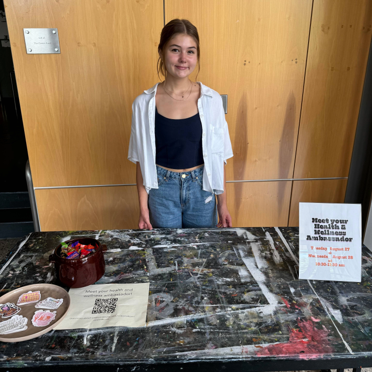 Photograph of Ella Teipen, Herron School of Art and Design's student health and wellness ambassador standing in front of a table covered with stickers and information about the wellness initiative