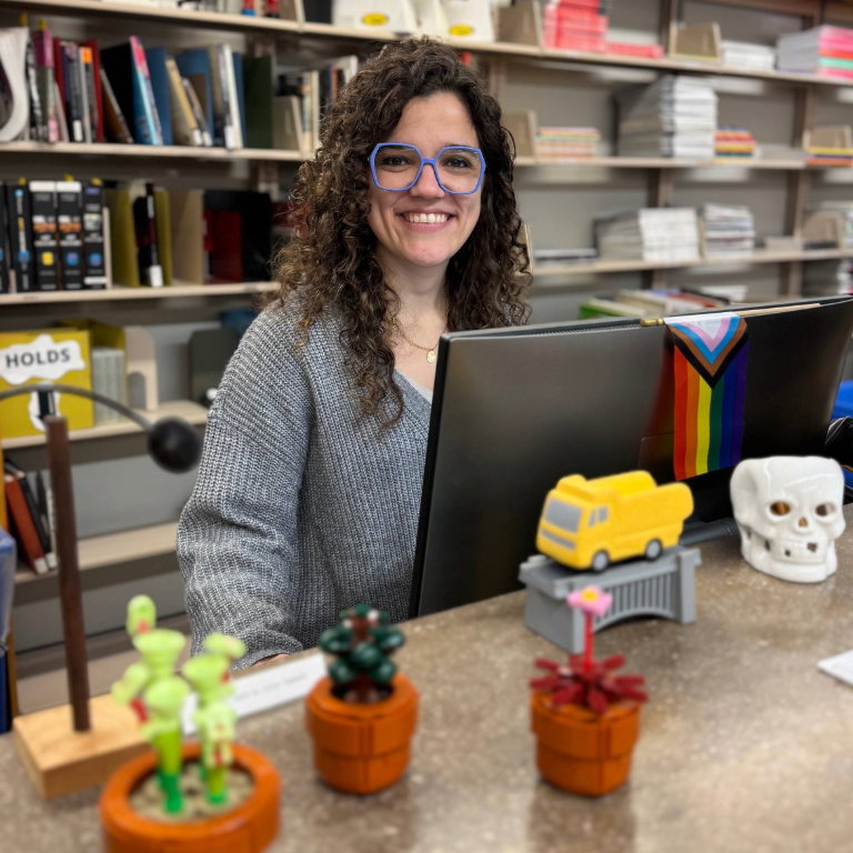 Herron Art and Design Librarian Jackie Huddle smiles behind the checkout counter in the Herron art library, which is covered in colorful figurines and knick-knacks